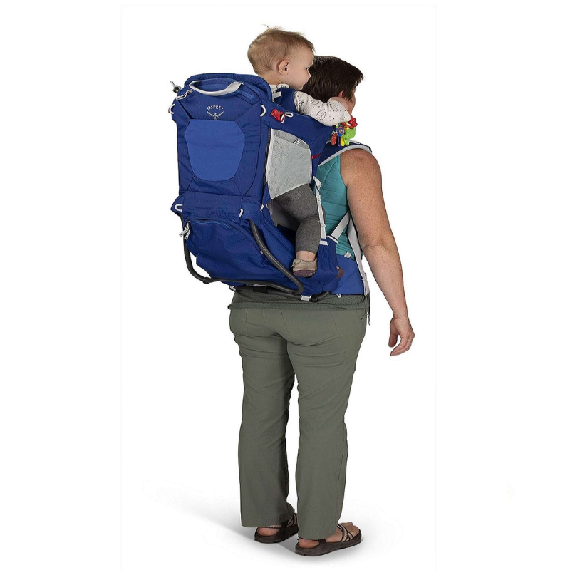 Un adulto vestido con pantalones color caqui y una camiseta sin mangas lleva a un niño pequeño en una mochila portabebé azul para senderismo, que recuerda la comodidad que proporciona la Mochila Portabebé Osprey Poco Child Carrier. El niño está sentado cómodamente, mirando hacia un lado, asegurado por el asiento incorporado y las correas de seguridad de la mochila.
