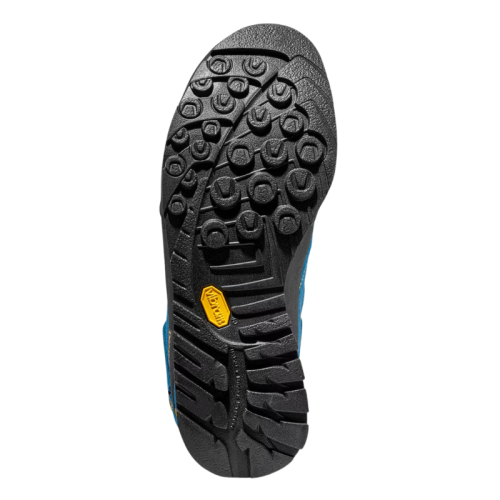 Primer plano de una suela de zapato negra con dibujo multipatrón similar a la Zapatilla de Aproximación La Sportiva Boulder X Mid GTX, que presenta ranuras circulares y lineales para tracción y un logotipo amarillo cerca del arco. La parte superior azul es parcialmente visible.