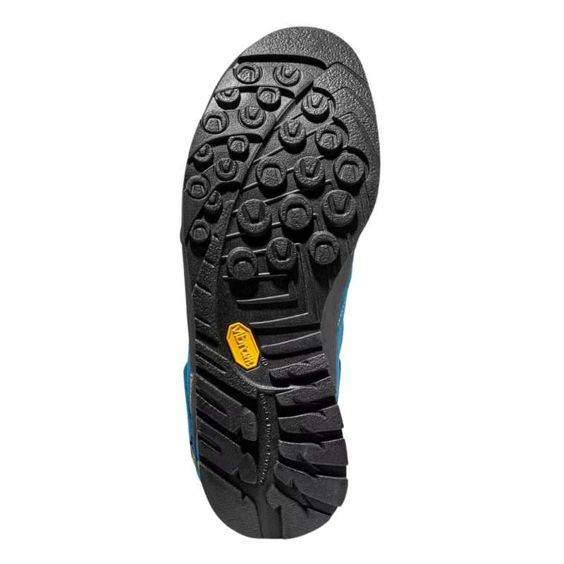 Primer plano de una suela de zapato negra con dibujo multipatrón similar a la Zapatilla de Aproximación La Sportiva Boulder X Mid GTX, que presenta ranuras circulares y lineales para tracción y un logotipo amarillo cerca del arco. La parte superior azul es parcialmente visible.