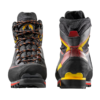 Un par de botas de montaña La Sportiva Karakorum Evo GTX, con detalles en negro, naranja y amarillo. La bota izquierda destaca los cordones en el lateral, mientras que la bota derecha muestra un llamativo tacón amarillo con elementos en rojo y amarillo.