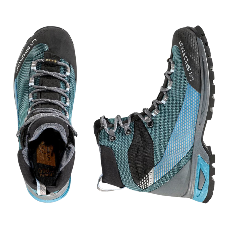 Un par de botas de senderismo similares a las La Sportiva Trango Trk GTX Mujer, con la bota izquierda mostrando su vista superior, cordones y plantilla "Ortho Hybrid". La bota derecha resalta un diseño azul grisáceo con detalles en negro y una suela resistente.