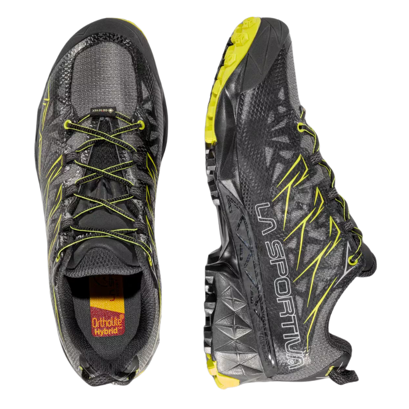 Las zapatillas de trail running en gris y negro, con detalles en amarillo y el nombre de la marca en el lateral, tienen suelas texturizadas para un mejor agarre. Los cordones se ven desde arriba, junto con una etiqueta "Ortholite Hybrid" en el interior, similar al estilo La Sportiva Akyra GTX.