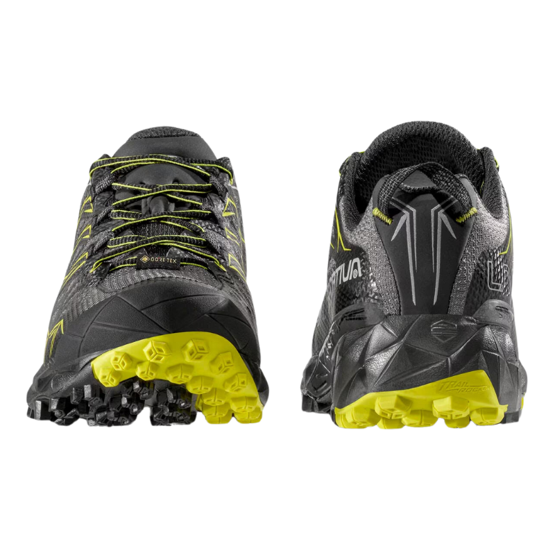 Las zapatillas de trail La Sportiva Akyra GTX en negro y amarillo se muestran de costado y desde atrás, con suelas resistentes y con tacos para mayor tracción. Tienen un diseño elegante con detalles reflectantes y el logotipo de la marca es visible tanto en el costado como en la parte posterior.