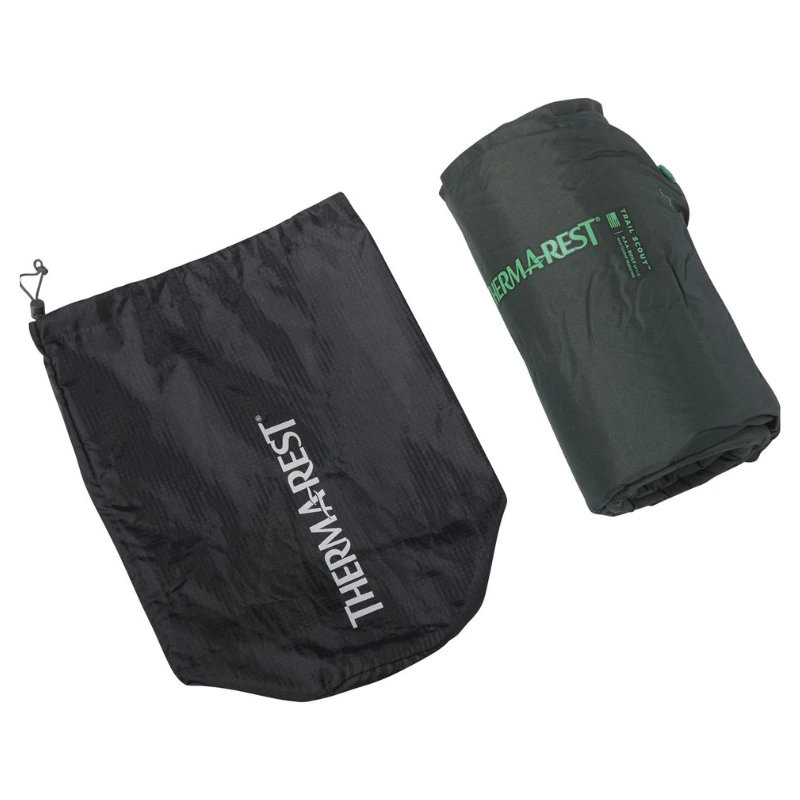 Una colchoneta enrollable para dormir Therm-a-Rest Trail Scout de color verde se encuentra junto a una bolsa de almacenamiento negra con cordón, ambas con el logotipo de Therm-a-Rest. Cerca de allí, una bolsa de dormir Pluma Therm-a-Rest Parsec 0F/-18C agrega el toque final al campamento sobre un fondo blanco liso.