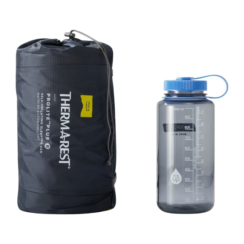 Una colchoneta para dormir compacta y de color gris oscuro, modelo Therm-a-Rest ProLite Plus, en una bolsa con cordón, se encuentra junto a una botella de agua Nalgene transparente con tapa azul, marcada con medidas de hasta 1000 ml.