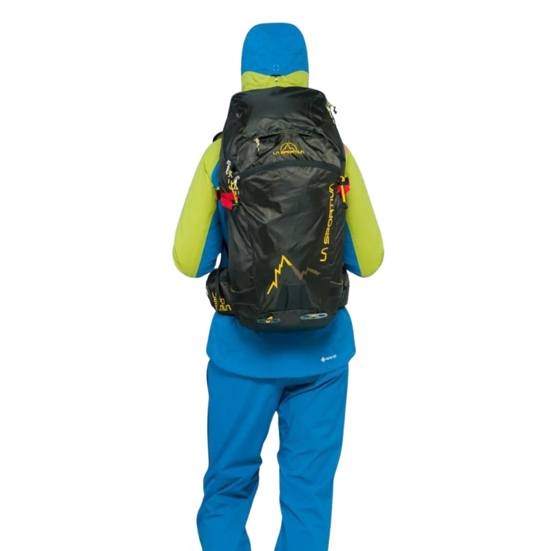 Una persona con ropa de montaña azul y verde mira hacia otro lado y lleva una mochila de trekking La Sportiva X-Cursion con logotipos amarillos. Con la capucha puesta, crea un aspecto listo para la aventura.
