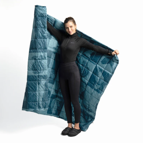 Una persona vestida con un traje negro y pantuflas sonríe mientras sostiene la gran Manta Térmica Multifuncional Atakama Outdoor Nalca azul con un patrón de cuadros, sobre un fondo blanco liso para mostrar la versatilidad y calidez de la manta.