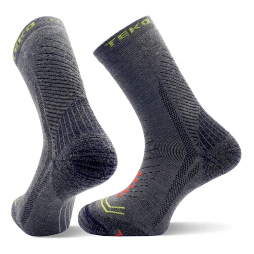Un par de calcetines de senderismo Calcetín Teko EcoHike Discovery Light Half Cushion 2.0 de color gris oscuro, con patrones texturizados y detalles en verde neón y naranja, se mantienen en posición vertical, resaltando las secciones del talón y la puntera.