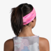 Una persona con cabello oscuro recogido en una cola de caballo mira hacia otro lado y lleva una cinta para el pelo Cinta Buff CoolNet UV Slim Thisy Multi, con un degradado rosa y naranja. Lleva una camiseta sin mangas con un patrón abstracto gris y rosa sobre un fondo blanco liso.