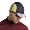 Una persona que viste una camiseta de ciclismo color granate y una gorra Jockey Buff Pack Cycle Glen Multi en negro, amarillo y blanco mira hacia abajo contra el fondo liso.
