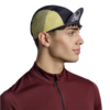Un hombre con una camiseta de color granate con cremallera lleva la gorra Jockey Buff Pack Cycle Lenir Night Blue, caracterizada por un estampado negro y amarillo con paneles de malla. El logotipo añade un toque deportivo mientras mira hacia la derecha sobre un fondo blanco sencillo, lo que resalta su aspecto moderno y activo.