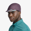 Una persona que lleva una gorra Jockey Buff Pack Cycle Xcross Multi y una chaqueta turquesa mira hacia la cámara. La gorra, con su estampado de rayas bajo la visera, complementa su pequeño pendiente de aro sobre un fondo blanco liso.