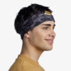 Una persona con cabello corto y oscuro que usa una diadema Bandana Buff Original EcoStretch Enhan Brindle Brown y un suéter mostaza sonríe y mira hacia un lado sobre un fondo blanco liso.