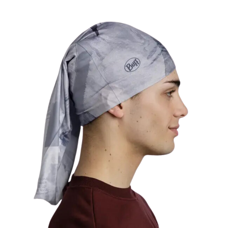 Una persona lleva un pañuelo gris largo en la cabeza con la leyenda "Bandana Buff Original EcoStretch Grafft 2.0 Multi (copia)", con el logotipo visible. Su pelo corto y oscuro se asoma mientras mira hacia la derecha de perfil, vestida con una camiseta granate sobre un fondo blanco liso.