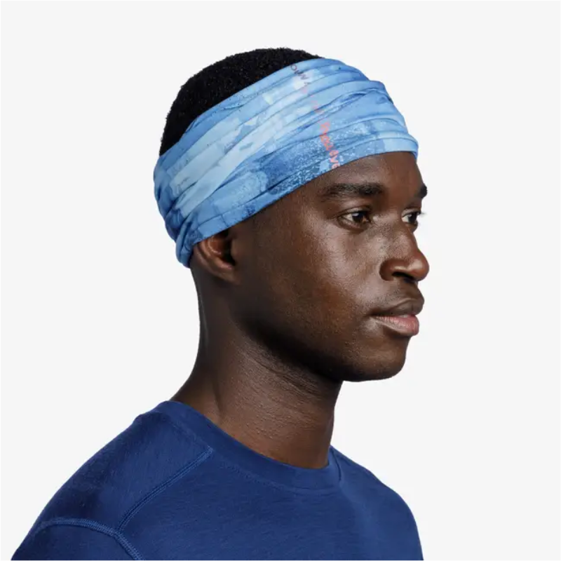 Una persona con una diadema estampada azul Bandana Buff Original EcoStretch Neski Multi (copia) y una camisa a juego mira de lado sobre un fondo liso.