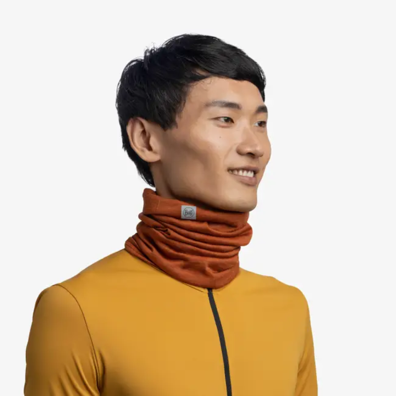 Una persona con un suéter mostaza de cuello alto sonríe mirando de reojo, luciendo una Bandana Buff Merino Lightweight Multistripes Maize. Este accesorio, con rayas de color maíz y naranja, le agrega estilo al fondo blanco liso.