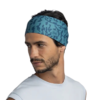 Un hombre con barba viste una camiseta sin mangas de color azul claro y una diadema Buff CoolNet UV Wide en Ater Teal, que revela su patrón de camuflaje mientras mira hacia la izquierda sobre un fondo blanco liso, exudando un estilo sencillo.