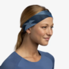 Una persona con el pelo largo recogido lleva la Cinta Buff CoolNet UV Wide Ater en color verde azulado, con un estampado geométrico. Lleva una camiseta azul y sonríe frente a un fondo liso, que encarna a la perfección la comodidad refrescante del material.