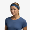 Una persona que lleva una cinta para la cabeza Cinta Buff CoolNet UV Slim Ahin Azure de color verde azulado y una camiseta de manga corta de color azul. Lleva el pelo castaño claro atado hacia atrás mientras mira directamente a la cámara con una expresión neutra sobre un fondo blanco liso.