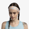 Una mujer con cabello trenzado, que lleva una diadema Cinta Buff CoolNet UV Slim Tasie Multi y una camiseta sin mangas azul claro, muestra su tatuaje floral mientras está de pie sobre un fondo liso, mirando hacia abajo.