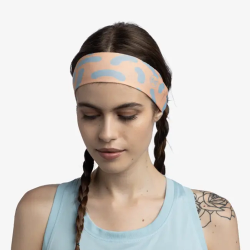 Una mujer con cabello trenzado, que lleva una diadema Cinta Buff CoolNet UV Slim Tasie Multi y una camiseta sin mangas azul claro, muestra su tatuaje floral mientras está de pie sobre un fondo liso, mirando hacia abajo.
