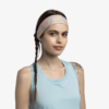 Una persona con trenza lleva una cinta para la cabeza Cinta Buff CoolNet UV Slim Tasie Multi y una camiseta sin mangas azul claro. Tiene un tatuaje en el hombro y una expresión neutra sobre un fondo blanco liso.