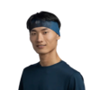Una persona que lleva una diadema Cinta Buff CoolNet UV Slim Tasie Multi y una camisa azul oscuro sonríe a la cámara sobre un fondo blanco liso.