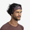 Una persona con cabello rizado lleva una cinta para el pelo estampada en tonos granate y negro, similar a la Cinta Buff CoolNet UV Slim Sishpink Fluor, combinada con una camisa granate. Mira hacia un lado con una expresión neutra sobre un fondo blanco liso.