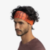 Con una diadema Cinta Buff Fastwick Ikshi Terracotta con un patrón sutil, que muestra parcialmente la marca "Buff", y vestida con una camisa granate, la persona de cabello rizado mira ligeramente hacia un lado contra un fondo claro y liso.