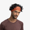 Un hombre de cabello rizado viste la diadema Cinta Buff Fastwick Ikshi Terracotta y una camisa granate, sonriendo levemente mientras mira directamente a la cámara contra un fondo blanco.