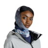 Una persona con una chaqueta gris y un pañuelo Bandana Buff ThermoNet Synth Blue mira hacia delante. El pañuelo le cubre la cabeza y el cuello en tonos azules, grises y blancos. La chaqueta está cerrada con cremallera sobre un fondo blanco liso.