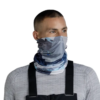 Una persona que viste una camisa gris de manga larga y tirantes negros luce una polaina para el cuello Bandana Buff ThermoNet Synth Blue, con diseños abstractos en tonos azul, gris y blanco.