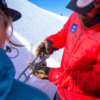 Una persona con una chaqueta roja utiliza la sierra de nieve MSR Beta para examinar una muestra de nieve mientras usa guantes y un sombrero, mientras alguien con una chaqueta azul observa la ladera nevada bajo el cielo azul claro.
