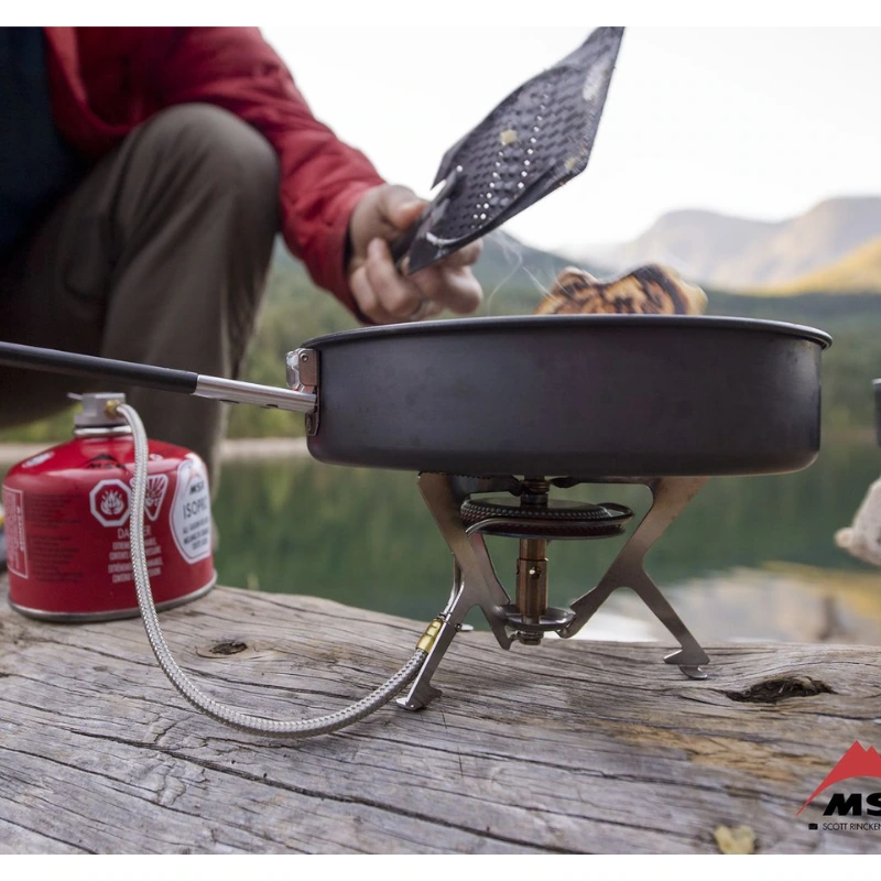 Una persona con una chaqueta roja da vuelta la comida en una sartén antiadherente MSR Quick Skillet sobre una estufa portátil sobre un tronco junto al lago, con un bote de combustible MSR contra un fondo de montañas y bosques.