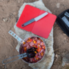 Vista desde arriba de una escena de preparación de comida para acampar sobre una roca. En ella se ve una olla con carne y verduras cortadas en cubitos y pinzas en el interior. Junto a ella, en la Base Estabilizadora MSR Trillium para Cocinillas (copia), hay una tabla de cortar roja con un cuchillo y, a la derecha, se ve parte de una bolsa negra.