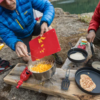 Dos personas cocinan al aire libre junto a un lago usando la Base Estabilizadora MSR Trillium para Cocinillas (copia) sobre un banco de madera. Una agrega pimientos amarillos picados a una olla, mientras que la otra prepara panqueques. Los utensilios y los ingredientes están esparcidos y ambos usan equipo para exteriores.