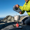 Una persona con una chaqueta verde vierte agua de una Bolsa de Hidratación Platypus Big Zip EVO en una olla sobre una estufa de camping portátil, frente a un paisaje de montaña rocosa y un cielo azul claro.