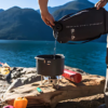Una persona vierte agua de una Bolsa de Agua MSR DromLite negra en una olla sobre una estufa de camping junto al lago. Cerca de allí, hay un kayak, pimientos morrones de colores, un encendedor y una bombona de combustible. Las montañas se alzan al fondo bajo el cielo azul claro.