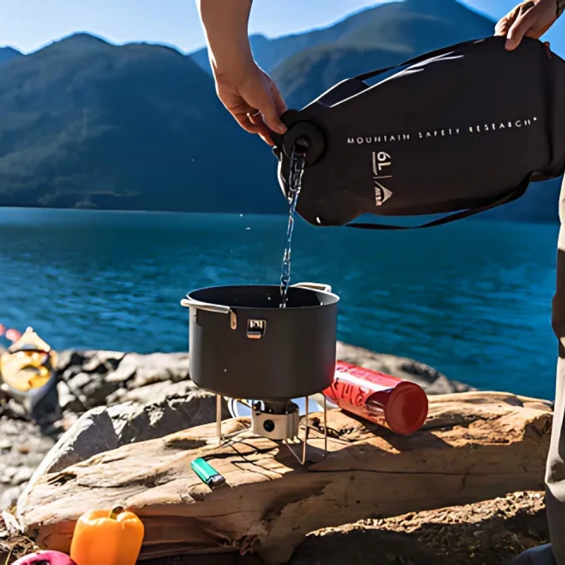 Una persona vierte agua de una Bolsa de Agua MSR DromLite negra en una olla sobre una estufa de camping junto al lago. Cerca de allí, hay un kayak, pimientos morrones de colores, un encendedor y una bombona de combustible. Las montañas se alzan al fondo bajo el cielo azul claro.