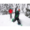 Un niño, equipado con Raquetas de Nieve MSR Shift Juvenil (copia) y un abrigo de invierno, camina juguetonamente por un bosque nevado delante de un adulto con una chaqueta roja, rodeado de árboles cubiertos de nieve.