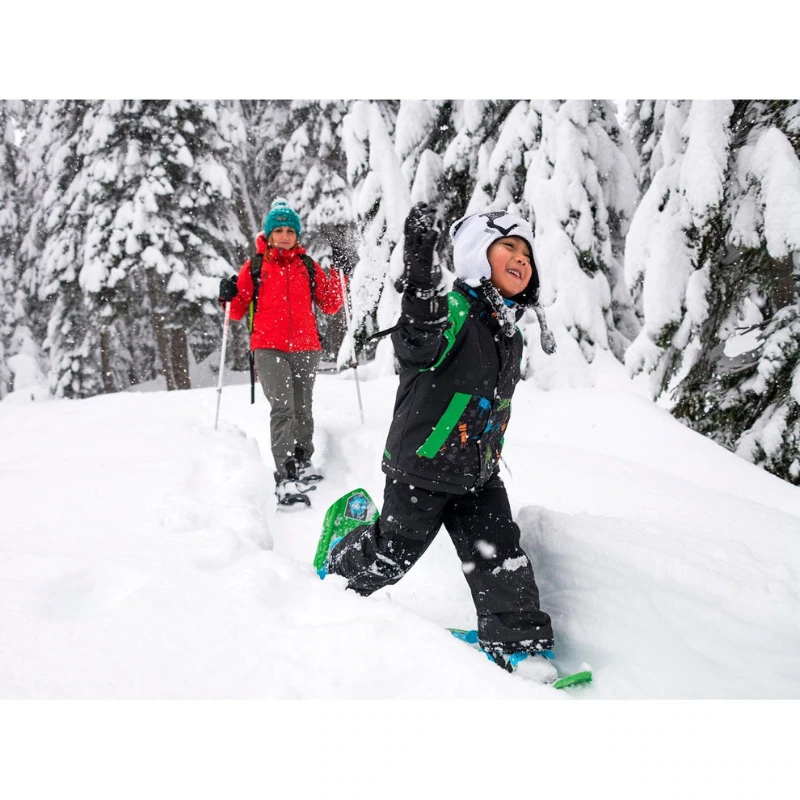 Un niño, equipado con Raquetas de Nieve MSR Shift Juvenil (copia) y un abrigo de invierno, camina juguetonamente por un bosque nevado delante de un adulto con una chaqueta roja, rodeado de árboles cubiertos de nieve.