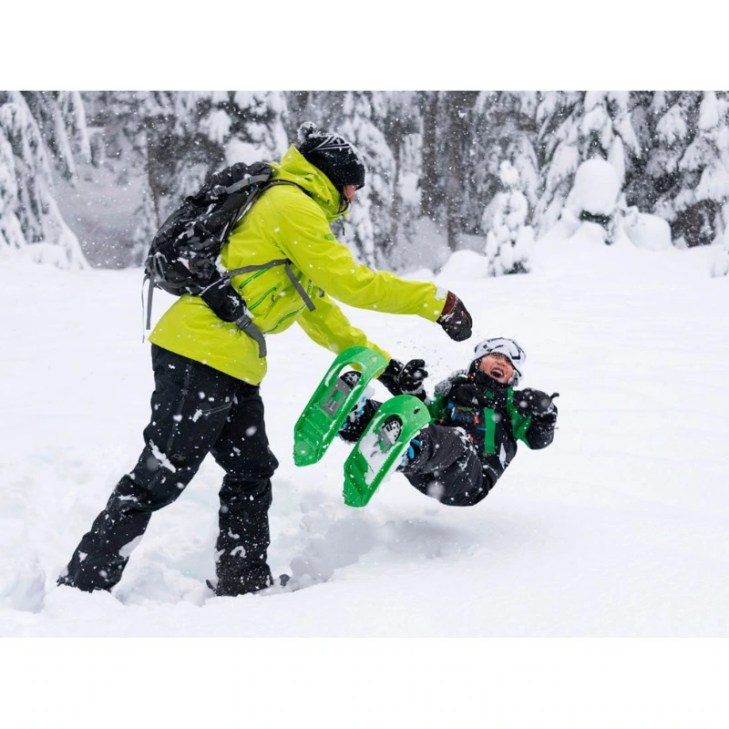 Un adulto con una chaqueta verde brillante y un niño con Raquetas de Nieve MSR Shift Juvenil (copia) juegan alegremente en el bosque nevado. Mientras el niño se balancea, ambos disfrutan de su aventura con raquetas de nieve entre los árboles cubiertos de nieve.