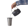 Una mano con una camisa a cuadros azul vierte agua de una tetera Olla MSR Titan de 0,8 l (copia) en una taza de acero inoxidable, ambos objetos aislados sobre un fondo blanco con un logotipo de la marca visible en la tetera.
