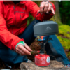 Una persona con una chaqueta roja usa una Olla MSR Titan Kettle 0.8L (copia) en una estufa de camping portátil con una bombona de gas para calentar agua al aire libre, rodeada de frondosos árboles verdes que sugieren un entorno forestal sereno.
