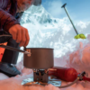 En un entorno nevado, alguien cocina con una estufa Cocinilla MSR WhisperLite Universal (copia), con una olla encima. Cerca hay un bidón de combustible rojo y una pala en la nieve, con montañas al fondo.
