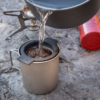 El agua fluye desde una olla con la Pinza para Ollas MSR PanHandler (copia) hacia una taza de camping de metal con un filtro de café, colocada sobre rocas. Una estufa portátil y un bidón de combustible rojo se suman a la robusta instalación para exteriores.