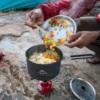 Una persona con una chaqueta roja cocina al aire libre en una estufa de camping portátil, usando su Tabla para Picar MSR Alpine Deluxe (copia) para preparar las comidas. Agrega verduras picadas de colores a una olla humeante, con una tienda de campaña visible en el fondo.