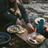 Una persona está al aire libre preparando comida, cortando un tomate en su Tabla para Picar MSR Alpine Deluxe (copia). Cerca hay platos con tortillas rellenas, hierbas frescas, carne en rodajas y mantequilla. Utensilios de cocina y sartenes están esparcidos en el suelo rocoso.