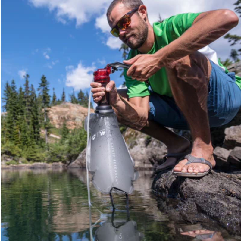 Un hombre con gafas de sol y una camiseta verde se agacha junto a un lago, utilizando su filtro de agua portátil Set de Cocina MSR Quick 2 (copia) para purificar el agua. Está rodeado de árboles y rocas bajo un cielo azul claro.