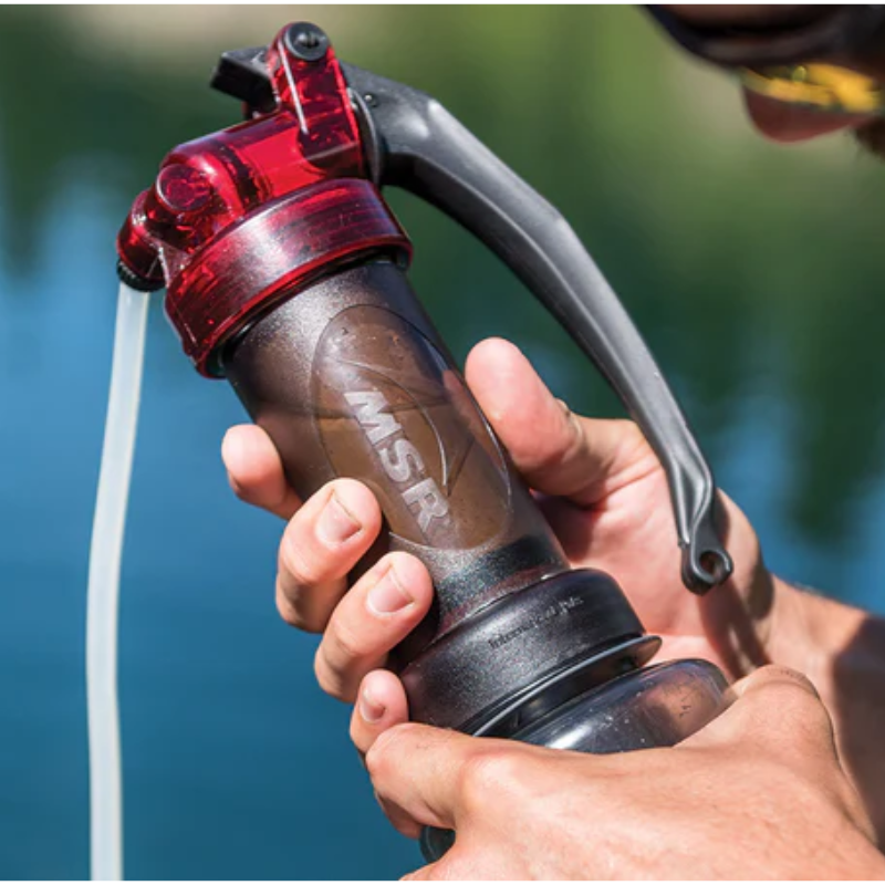 Una persona sostiene una bomba de filtro de agua portátil con detalles en negro y rojo cerca del agua, sacándola a través de un tubo blanco. El Set de Cocina MSR Quick 2 (copia) que se encuentra cerca garantiza que estén listos para cualquier aventura de cocina al aire libre.